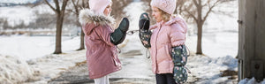 Two young female children play patty-cake on a snowy path in winter, wearing Stonz Winter Mitts & Snow Boots in Evergreen and Woodland Print.