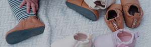 Infant's feet visible sitting on a cream blanket, surrounded by Stonz vegan baby shoes in Tan, Ivory & Pink colors.