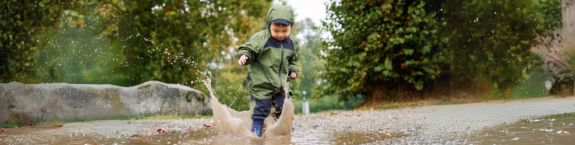 Child standing in forested area, smiling, head tipped ear to shoulder, wearing Stonz Rain Suit in Dusty Rose.