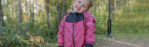 Child standing in forested area, smiling, head tipped ear to shoulder, wearing Stonz Rain Suit in Dusty Rose.