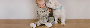 Little baby wearing a Stonz olive green gingham print sun suit and camel cruiser sneakers getting kissed by a cute white dog