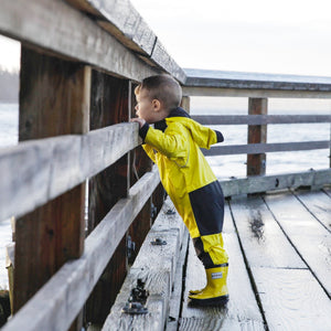 Stonz Rain Suit and matching Rain Boots in Yellow, worn by an infant peering out at the water from a deck.